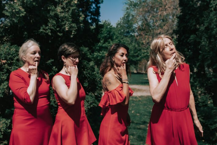 The all-female cast of Skin Songs in their red dresses, each with one finger touched to their bottom lip. They are in a park with evergreen trees in the background.