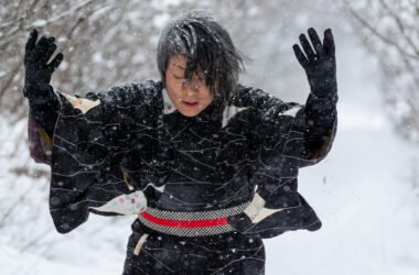 A photograph of Takako, a Japanese Canadian woman with short grey-streaked black hair, wearing a black kimono. Her head is bent down and her arms are raised up, gloved hands open. She is outside on a snowy day with bare trees behind her.