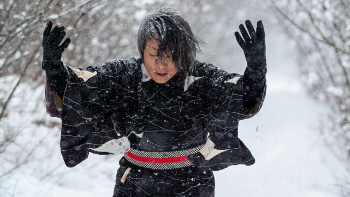 A photograph of Takako, a Japanese Canadian woman with short grey-streaked black hair, wearing a black kimono. Her head is bent down and her arms are raised up, gloved hands open. She is outside on a snowy day with bare trees behind her.